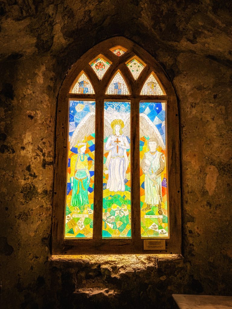 Ein buntes Glasfenster mit drei Figuren leuchtet in einer dunklen steinernen Nische.