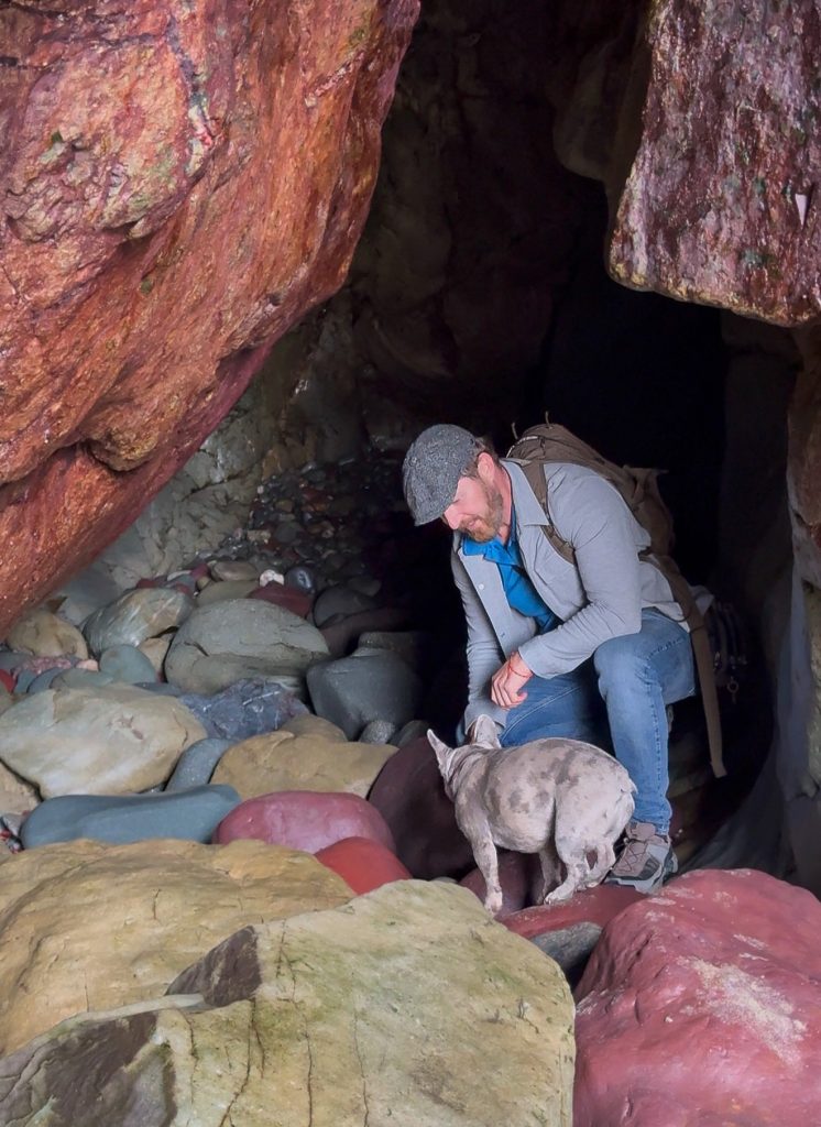 Heiko kniet mit Mila in einer Felshöhle bei Caerfai Bay zwischen großen, bunten Kieselsteinen.