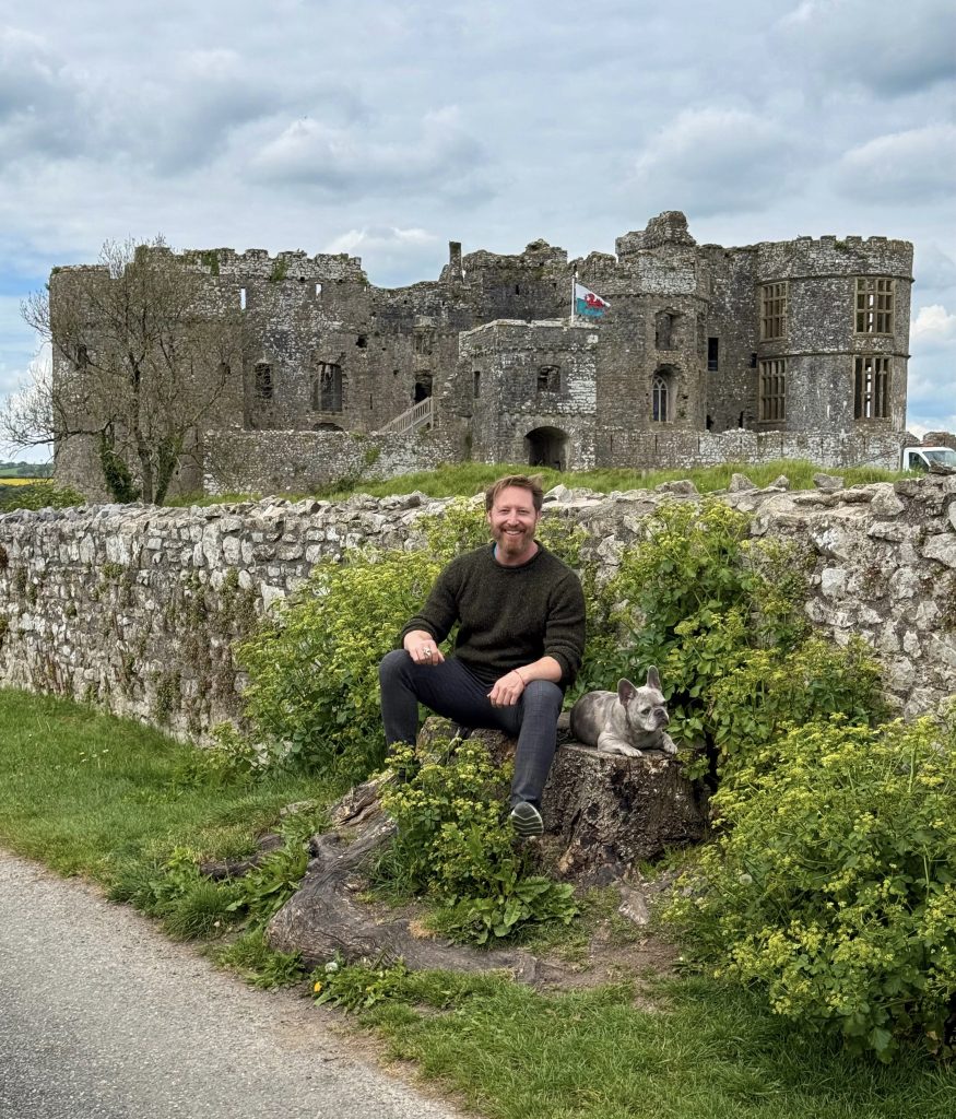 Heiko sitzt mit Mila vor den Mauern von Carew Castle; auf der Burg weht die walisische Flagge.