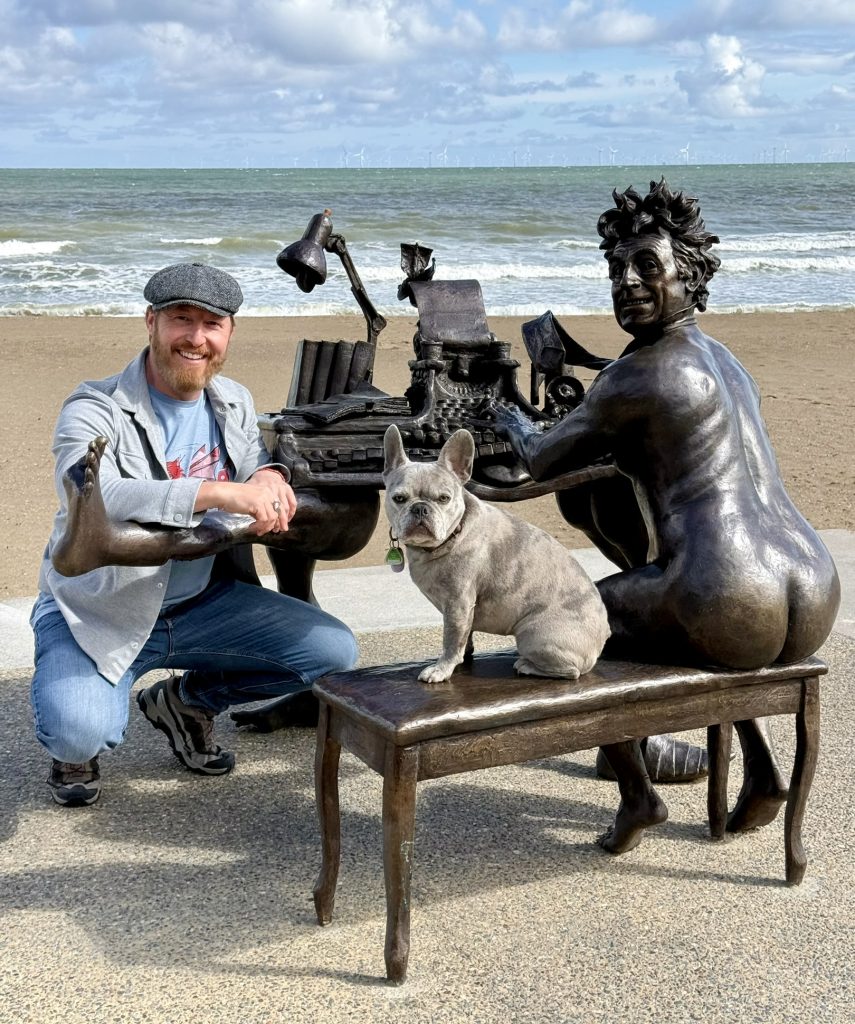 Heiko und Mila sitzen neben der Terry Jones Bronzeskulptur am Strand; dahinter sind das Meer und Windräder zu sehen.