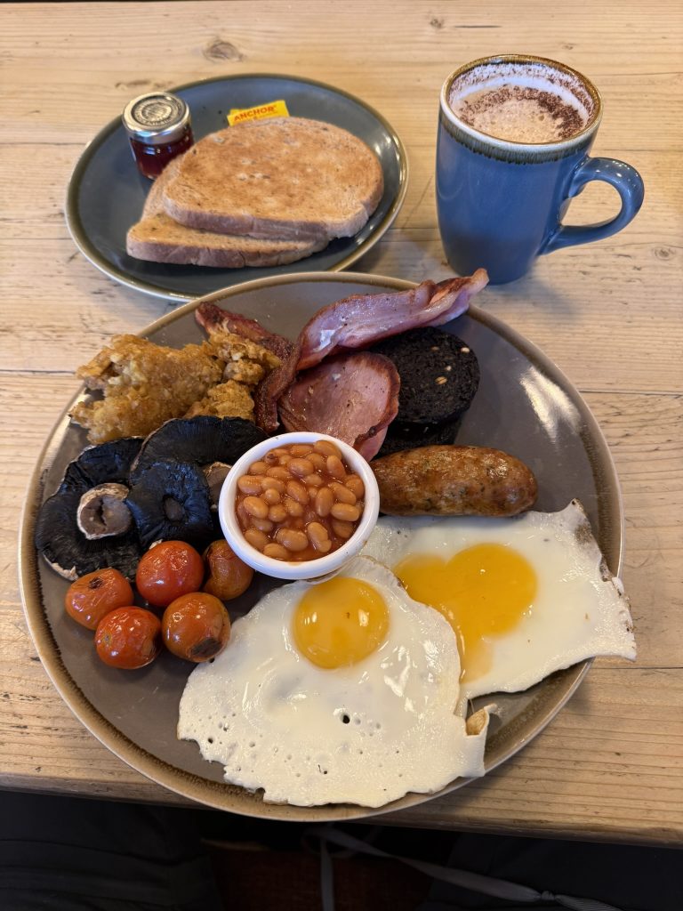 Ein herzhaftes Frühstück mit Spiegeleiern, Würstchen und Bohnen. Daneben stehen Kaffee und Toast.