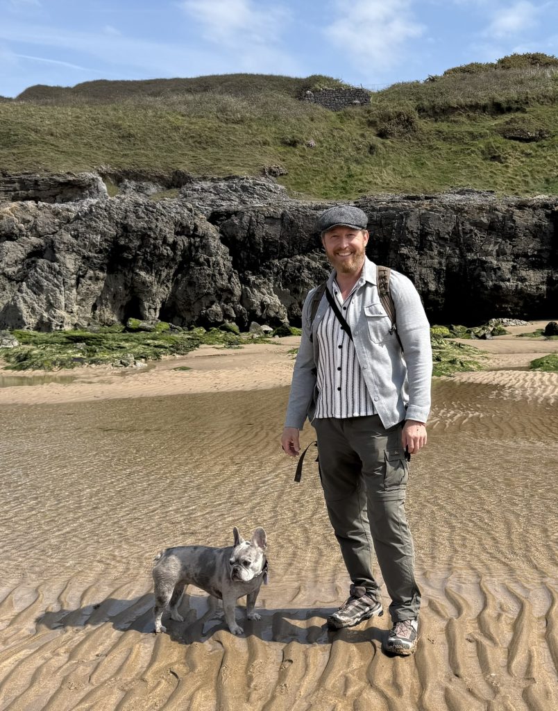 Heiko steht mit Mila auf dem feuchten Sand an der Küste der Gower-Halbinsel. Hinter ihnen liegen dunkle Felsen.