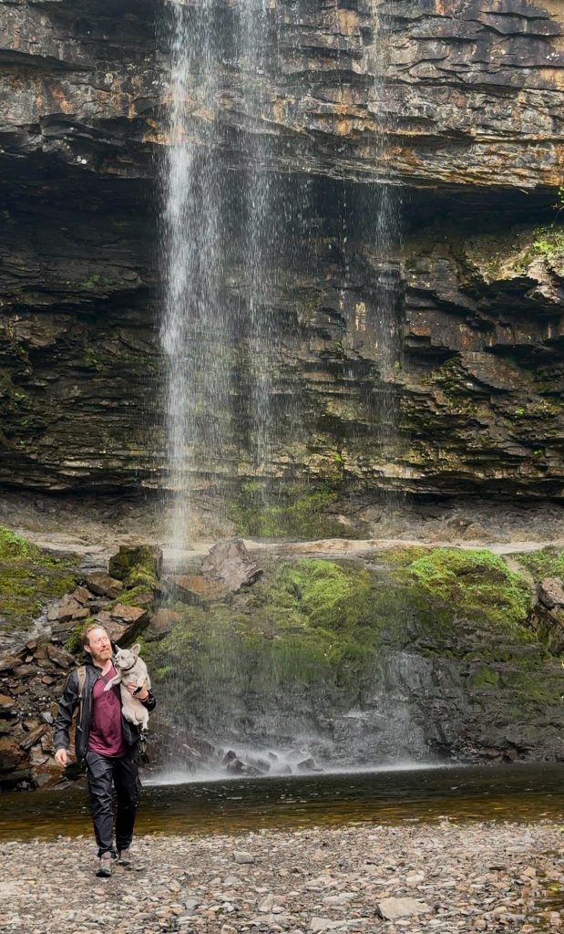 Heiko trägt Mila durch das flache Wasser vor einem Wasserfall.
