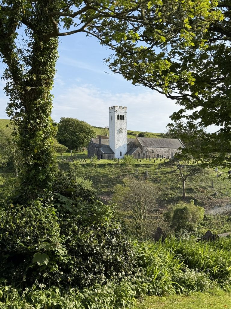 Eine kleine steinerne Kirche mit weißem Turm und vielen Grabsteinen steht mitten im Grün.
