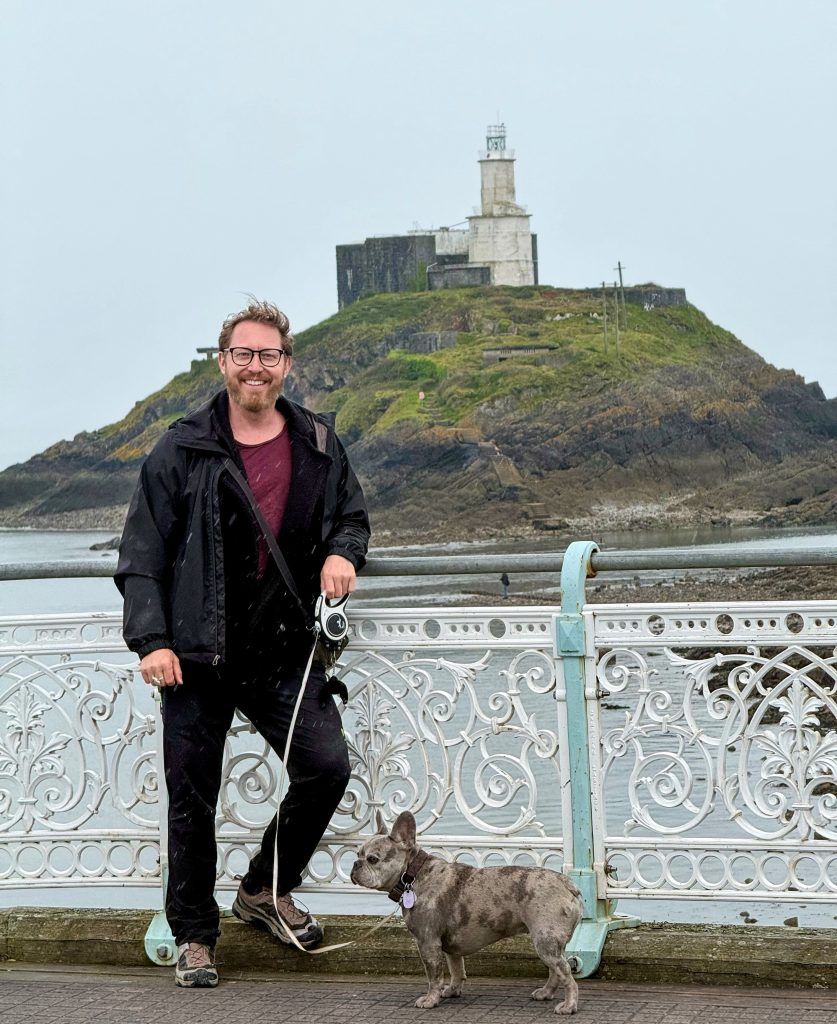 Heiko und Mila stehen an einem weißen Geländer in Mumbles; im Hintergrund steht ein Leuchtturm auf einer kleinen, grünen Insel.