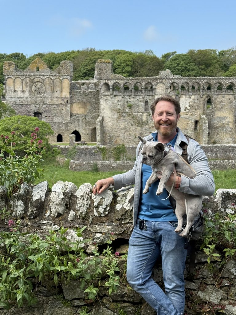 Heiko hält Mila im Arm und steht mit ihr vor einer hüfthohen Mauer. Hinter ihn steht die Ruine eines burgähnlichen Bischofspalastes.