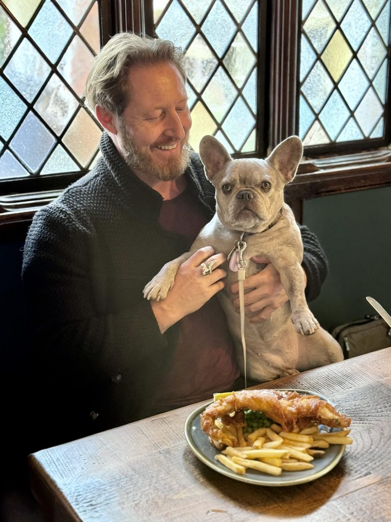 Heiko sitzt mit Mila an einem Tisch im Pub; vor ihnen steht ein Teller mit Fish and Chips.