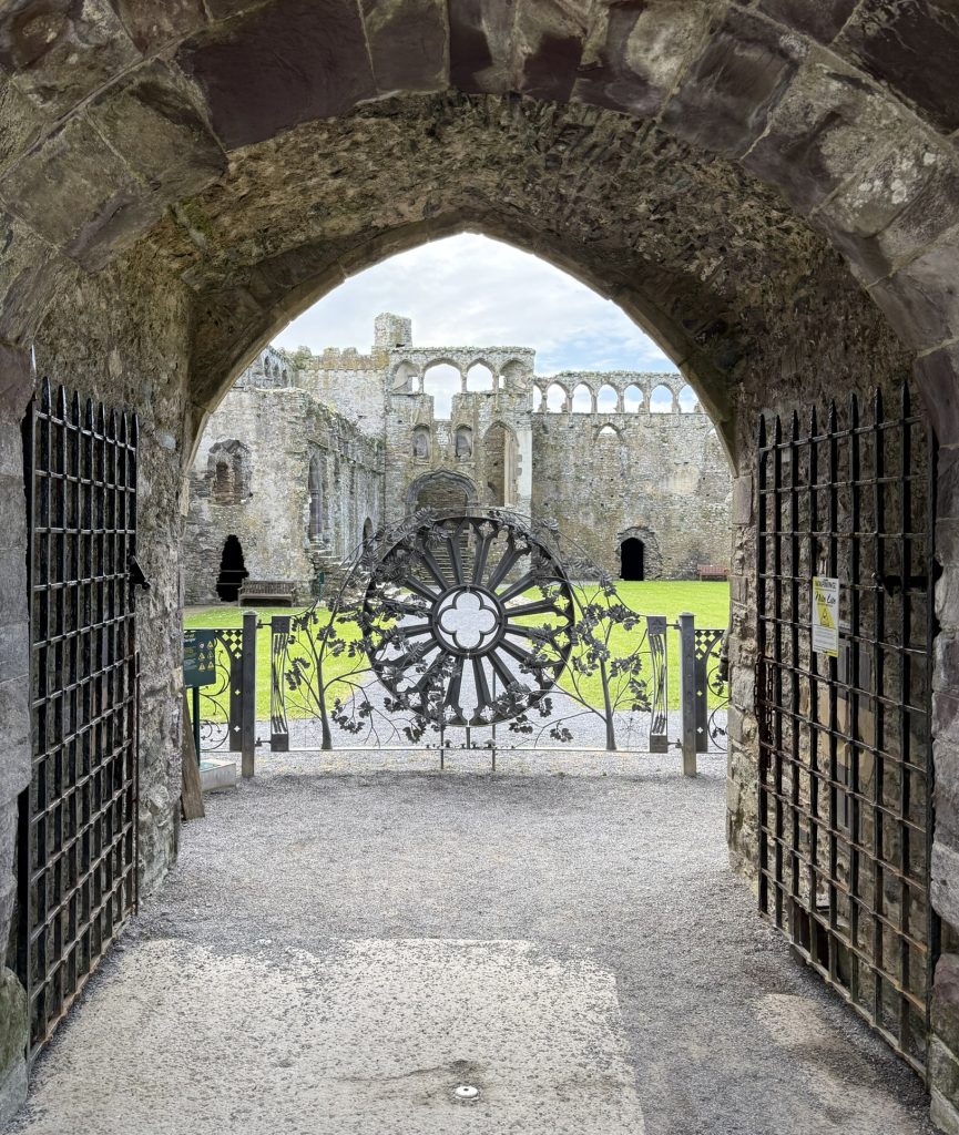 Durch ein steinernes Tor fällt der Blick auf die Ruinen des Bischofspalastes und ein kunstvolles Metallrad.